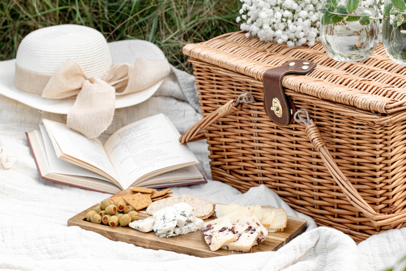 A wicker picnic hamper beside a sun hat, an open book and a board of cheese and crackers